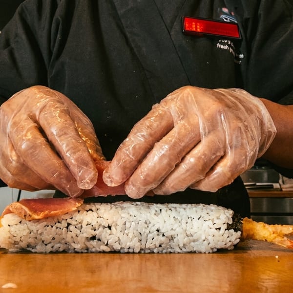 Chef's gloved hands placing tuna on a sushi roll with tempura shrimp on a wooden counter.